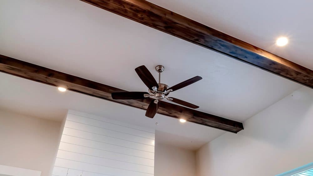 Recessed lighting in a room with high ceilings with two darker wooden beams running perpendicular and surrounding a ceiling fan