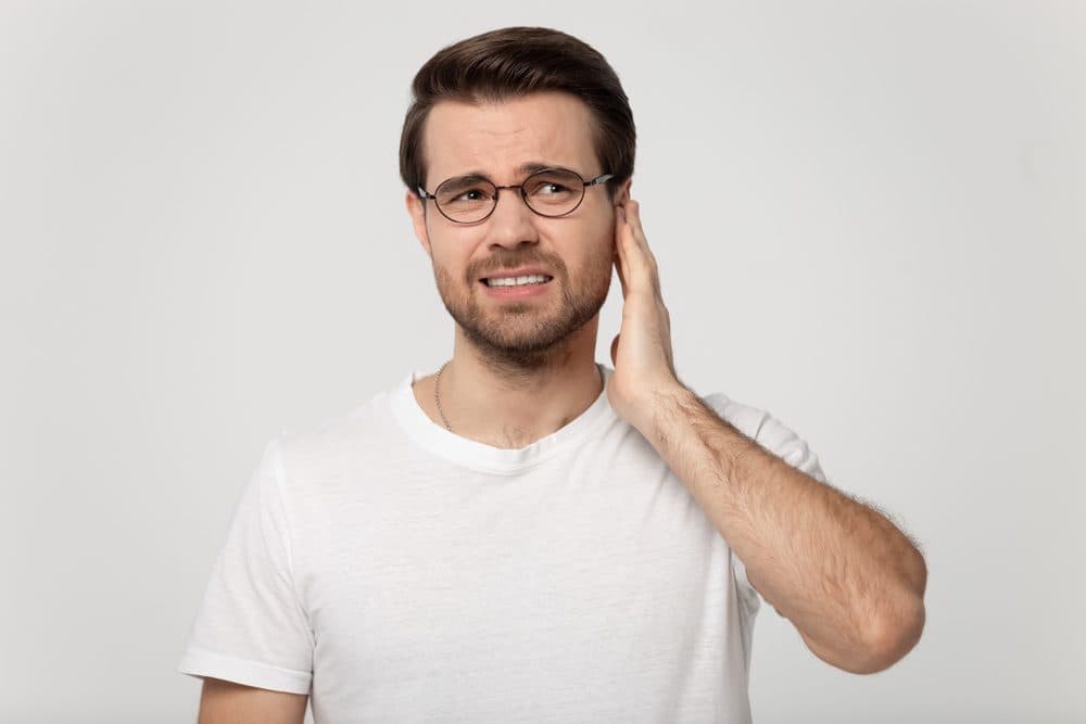A white male wearing glasses and a t-shirt with his left hand held up to his ear like the ear is in pain