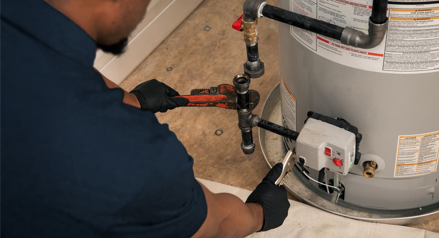 A Horizon plumber wearing black gloves uses a pipe wrench to repair the gas line and control valve assembly on a residential water heater tank.