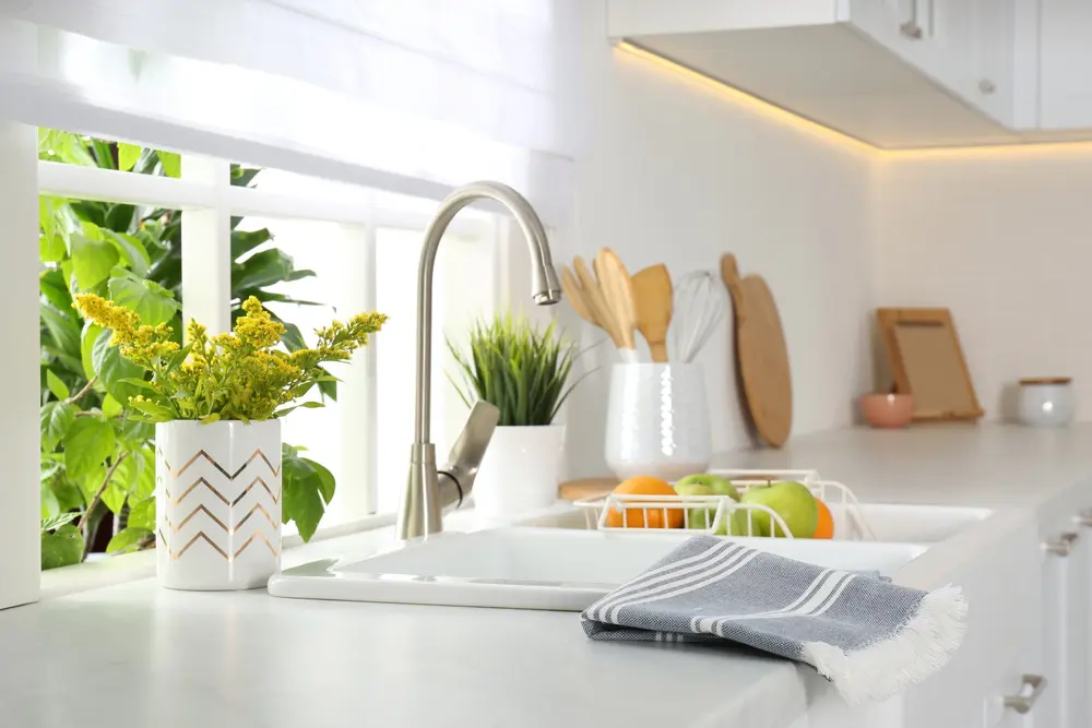Focused on a kitchen sink in an all-white, modern kitchen with green plants surrounding the sink and a tea towel sitting on the counter top