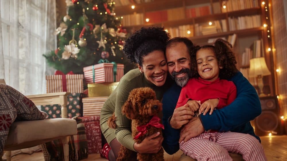 A mother, father, daughter, and dog, all smiling and hugging underneath a Christmas tree with warm lights in the background